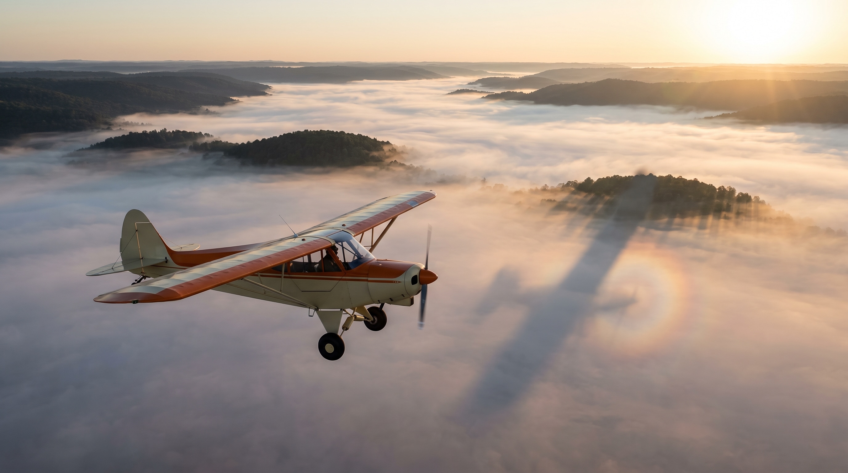 Ultraleichtflugzeug im Sonnenaufgang über einem Nebelmeer, aus dem bewaldete Hügel ragen; der Schatten des Flugzeugs liegt auf dem Nebel